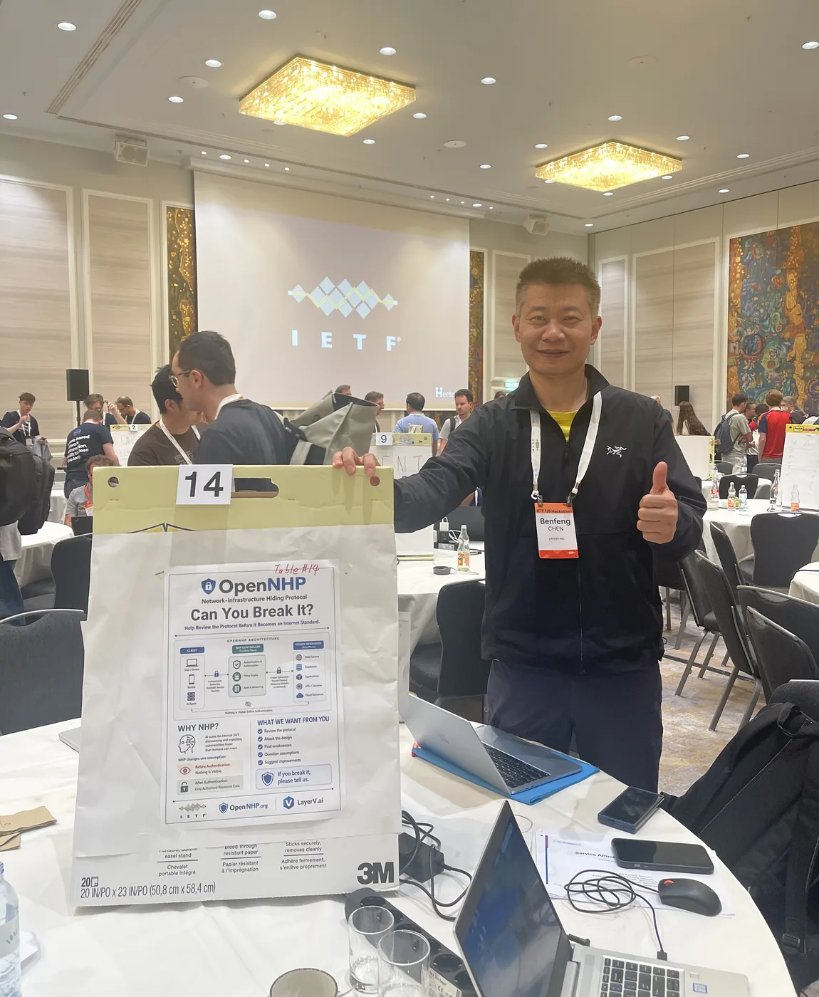 Benfeng Chen at OpenNHP's Table #14 at the IETF 126 Hackathon in Vienna, giving a thumbs up beside the "OpenNHP — Can You Break It?" poster, with the packed hackathon floor and IETF screen behind him
